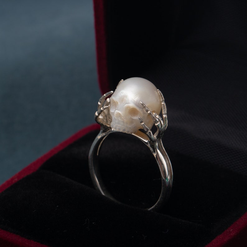 May include: A silver ring with a white pearl skull design. The ring is in a red velvet box.