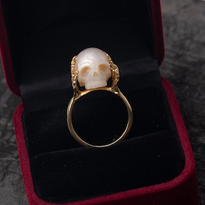 May include: A gold ring with a white skull made of mother of pearl. The ring is in a red velvet box.