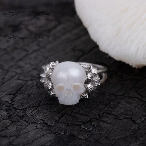 May include: A silver ring with a white pearl skull design. The ring is set against a black background.