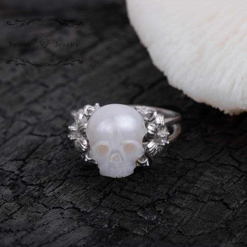 May include: A silver ring with a white pearl skull design. The ring is set against a black background.