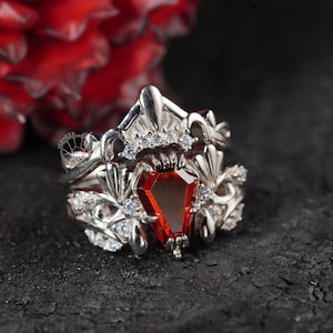 Puede incluir: Un anillo de plata con una piedra roja en forma de ataúd. El anillo está adornado con detalles intrincados y pequeñas piedras blancas.