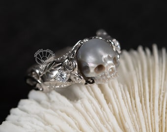 Puede incluir: Un anillo de plata con un diseño de calavera de perla. El anillo está engastado con pequeños diamantes y tiene un motivo floral.