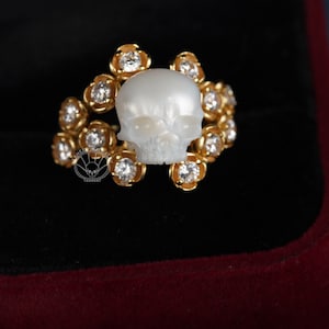 Op de afbeelding: Een gouden ring met een witte schedel gemaakt van parelmoer. De schedel wordt omringd door een halo van kleine, heldere diamanten.