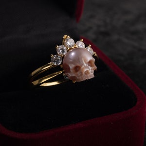 Op de afbeelding: Een gouden ring met een roze schedel van parelmoer en een kroon van heldere zirkonia stenen.