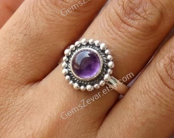 May include: A silver ring featuring a round, deep purple gemstone at the center, surrounded by a beaded design. The ring is set on a hand, showcasing the intricate details of the jewelry.
