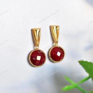 Op de afbeelding: Een paar goudkleurige oorbellen met een driehoekige bovenkant en een ronde, facetgeslepen, dieprode edelsteen. De oorbellen worden gepresenteerd op een witte achtergrond met een groen blad in de hoek.