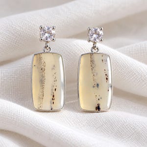 May include: A pair of drop earrings featuring rectangular, cream-coloured stones with dark, dendritic inclusions. Each stone is set in a silver-toned frame and suspended from a single, round, clear faceted stone stud with a four-prong setting.