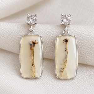 May include: A pair of silver drop earrings featuring rectangular, translucent, cream-coloured stones with dark, fern-like dendrite inclusions. Each earring is topped with a single, round, clear, faceted gemstone set in a four-prong silver mount.