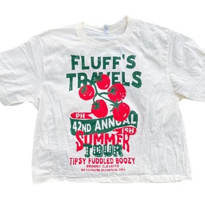 May include: White t-shirt with a crew neck and short sleeves. The front features a graphic with the text "FLUFF'S TRAVELS" in green, a cluster of red tomatoes, and the text "42ND ANNUAL SUMMER TOUR" in red. The shirt also has the text "TIPSY FUDDLED BOOZY".