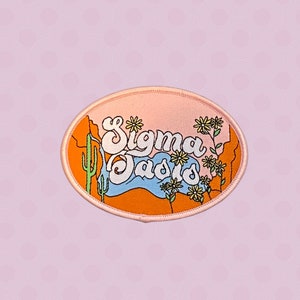 May include: A pink and orange embroidered patch with the text "Sigma Sads" in a retro font. The patch features a desert landscape with cacti and flowers.
