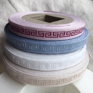 May include: A stack of spools of decorative ribbon in various colors: light pink, blue, white, and beige. Each ribbon features a repeating geometric pattern in a contrasting color. The spools are stacked on top of each other.