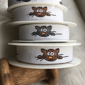 Peut inclure: Quatre bobines de ruban blanc avec un motif répété de chats de dessin animé. Les chats sont tournés vers l'avant, les yeux ouverts et ont des fourrures de couleurs différentes. Le ruban est enroulé autour des bobines et est prêt à être utilisé pour des projets d'artisanat.