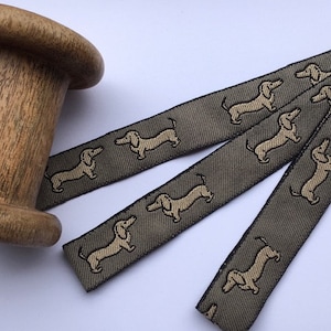 Op de afbeelding: Drie spoelen grijs lint met een beige teckelprint. Het lint is gevouwen en op een wit oppervlak gelegd.