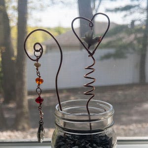 Può includere: Un barattolo decorativo con due ornamenti in filo di rame. Un ornamento presenta una forma a cuore con un piccolo uccello bianco, l'altro ha un design a spirale con perline pendenti nei colori arancione, rosso e trasparente. Il barattolo è riempito di ciottoli scuri.