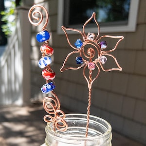 May include: Two copper wire garden stakes. One stake features a spiral top and a vertical row of blue, red, and white patterned glass beads. The other stake features a six-petaled flower shape formed from copper wire, accented with small, multi-colored faceted crystal beads.