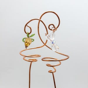 Può includere: Tappo per bottiglia di vino in filo di rame con un design decorativo. Presenta un ciondolo di perline gialle e verdi e un ciondolo di cristallo trasparente. Il filo è modellato in eleganti spirali e anelli, creando un aspetto unico e artistico.