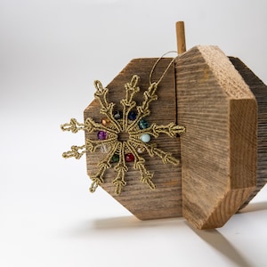 May include: A gold-colored snowflake ornament with colorful beads is displayed on a rustic wooden block. The snowflake has intricate details and a delicate appearance. The wooden block has a natural, weathered look, adding a touch of warmth and texture to the scene.
