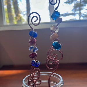 Può includere: Due paletti per piante fatti a mano in filo di rame con perline di vetro colorate. Ogni paletto presenta una parte superiore a spirale e un fiore decorativo alla base. Le perline sono blu, rosse, bianche e gialle. I paletti sono esposti in un barattolo di vetro trasparente.