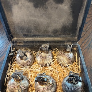 May include: A dark wooden box containing six baseball-shaped objects, each with red stitching and metal detailing, resembling grenades. They rest on straw. The box's interior has a distressed appearance.