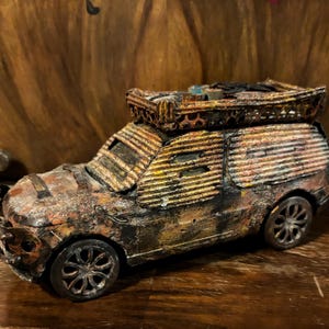 Könnte beinhalten: Ein rostiges, verwittertes und stark modifiziertes Spielzeugauto mit einem provisorischen Dachgepäckträger. Das Auto ist in Braun-, Orange- und Goldtönen lackiert, mit einer schwarzen Basis. Der Dachgepäckträger ist aus Metall und hat einen Korb oben drauf.
