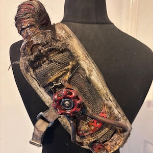 Puede incluir: Un bolso de hombro único y hecho a mano con una estética steampunk. El bolso presenta una combinación de tejidos texturizados, detalles metálicos y una prominente válvula roja. El diseño incluye una correa y se muestra en un maniquí negro.