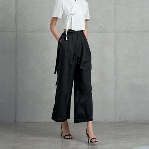 Puede incluir: Una blusa blanca recortada con un lazo en la parte delantera y mangas cortas. La blusa se combina con pantalones negros de pierna ancha.