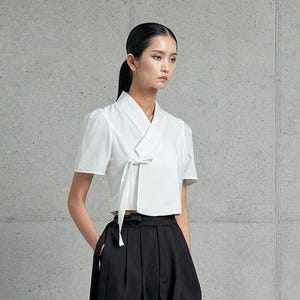 May include: A white short-sleeve top with a wrap-style neckline and tie closure, paired with high-waisted, wide-leg black pants. The outfit is completed with black heeled sandals. The ensemble is set against a neutral gray background.