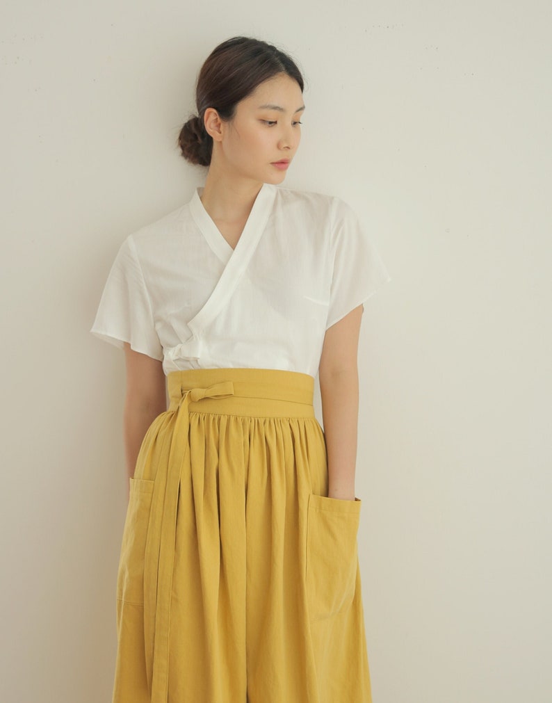 May include: A woman wearing a white, short-sleeved, V-neck top with a yellow skirt. The skirt has two pockets and a matching yellow sash tied at the waist.