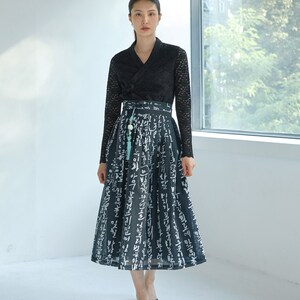May include: A black long-sleeved lace top with a V-neckline paired with a black midi skirt featuring a white Korean calligraphy print. The skirt has a high waistband with a decorative light blue tassel accessory hanging from the front side.