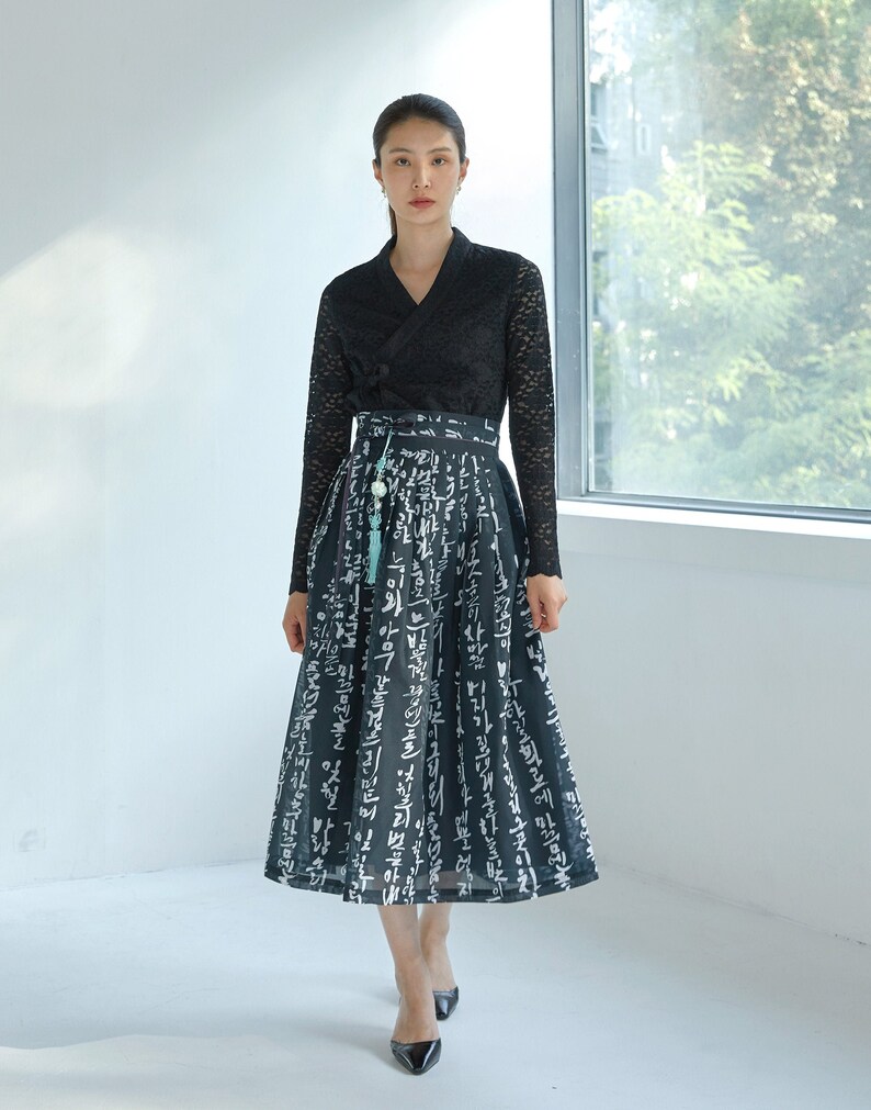 May include: A black long-sleeved lace top with a V-neckline paired with a black midi skirt featuring a white Korean calligraphy print. The skirt has a high waistband with a decorative light blue tassel accessory hanging from the front side.