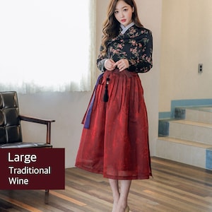 Może przedstawiać: Kobieta w tradycyjnej koreańskiej sukni hanbok. G&oacute;ra jest czarna z kwiatowym nadrukiem, a sp&oacute;dnica jest czerwona. Suknia jest związana niebieską wstążką. Kobieta stoi na drewnianej podłodze.