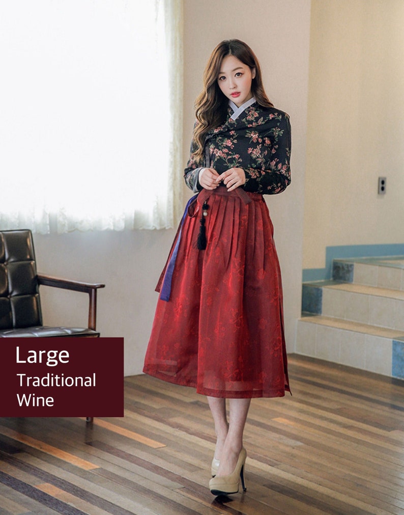 Może przedstawiać: Kobieta w tradycyjnej koreańskiej sukni hanbok. G&oacute;ra jest czarna z kwiatowym nadrukiem, a sp&oacute;dnica jest czerwona. Suknia jest związana niebieską wstążką. Kobieta stoi na drewnianej podłodze.