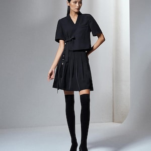 May include: A black short-sleeved wrap top with a side tie closure paired with a black pleated mini skirt. The outfit includes black over-the-knee socks and black pointed-toe high-heeled shoes, creating a monochromatic ensemble.