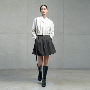 May include: A cropped off-white long-sleeve jacket with a high collar and a front zip, paired with a dark grey pleated mini skirt featuring a side belt detail and black knee-high boots. The ensemble creates a structured and layered appearance.