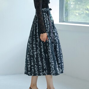 May include: A black long-sleeved lace top paired with a high-waisted, calf-length skirt featuring a black base with white Korean calligraphy print. The skirt has a wide waistband with a tie closure and a small decorative tassel accessory.