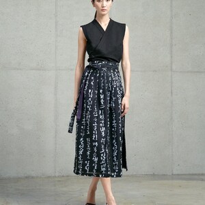 May include: A sleeveless black wrap top with a V-neckline paired with a black midi skirt featuring a white calligraphic print and a matching tie-waist belt. The skirt has a side slit and the ensemble is worn with black open-toe heeled sandals.