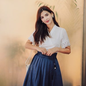 Puede incluir: Una mujer con una blusa blanca con ribetes de encaje y una falda plisada azul marino con un borla. Est&aacute; de pie frente a una pared de vidrio con una planta detr&aacute;s de ella.