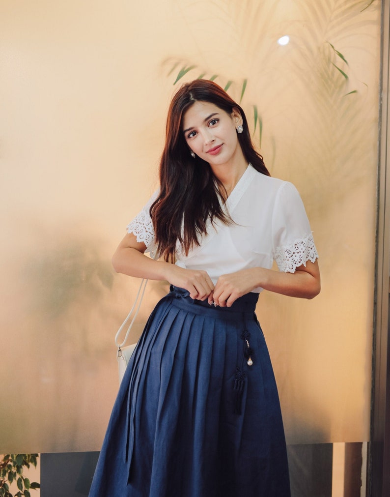 Puede incluir: Una mujer con una blusa blanca con ribetes de encaje y una falda plisada azul marino con un borla. Est&aacute; de pie frente a una pared de vidrio con una planta detr&aacute;s de ella.