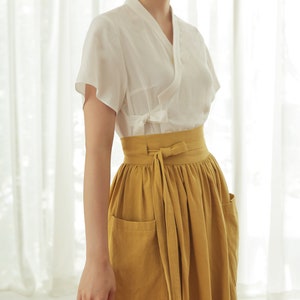 May include: A white short-sleeved blouse with a V-neck and a tie at the waist, worn with a mustard yellow skirt with pockets and a tie at the waist.