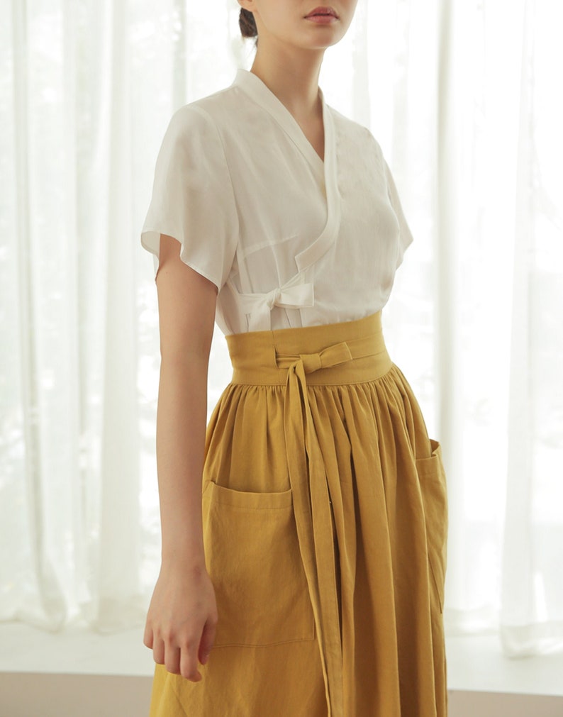 May include: A white short-sleeved blouse with a V-neck and a tie at the waist, worn with a mustard yellow skirt with pockets and a tie at the waist.