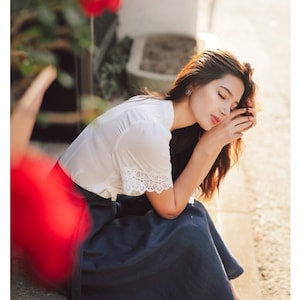 Puede incluir: Una mujer con una blusa blanca con ribetes de encaje y una falda azul oscuro se sienta en un escal&oacute;n de piedra con la cabeza apoyada en la mano.