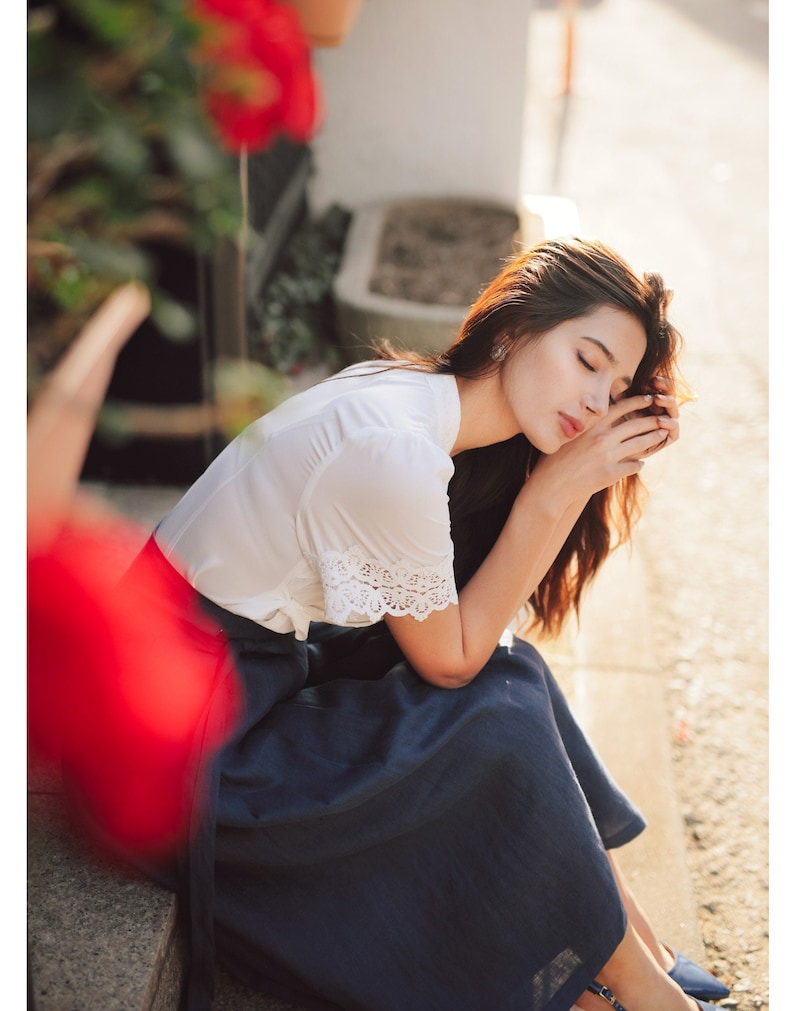 Puede incluir: Una mujer con una blusa blanca con ribetes de encaje y una falda azul oscuro se sienta en un escal&oacute;n de piedra con la cabeza apoyada en la mano.