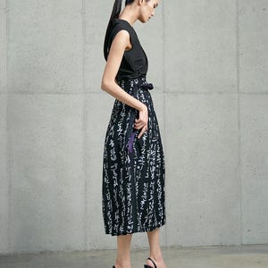May include: A sleeveless black wrap top paired with a high-waisted midi skirt featuring a black background and white calligraphic text print. The skirt includes a side tie closure and a purple accent detail visible at the waistline.