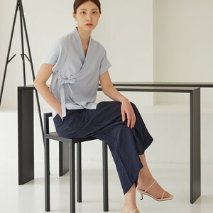 May include: A woman wearing a light blue linen wrap top with a navy blue wide-leg pant and cream-colored heeled sandals. She is sitting on a black metal stool.