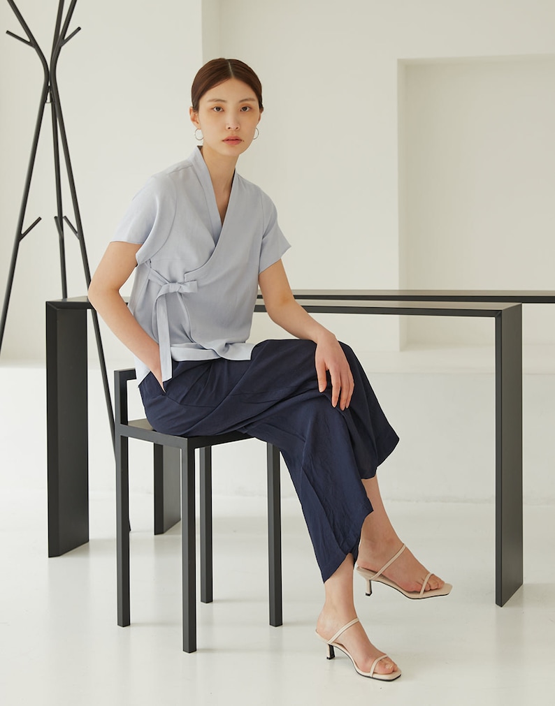 May include: A woman wearing a light blue linen wrap top with a navy blue wide-leg pant and cream-colored heeled sandals. She is sitting on a black metal stool.