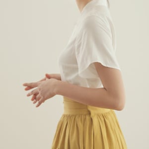 May include: A woman wearing a white short-sleeved blouse and a yellow midi skirt with a pocket. The skirt has a wide waistband.