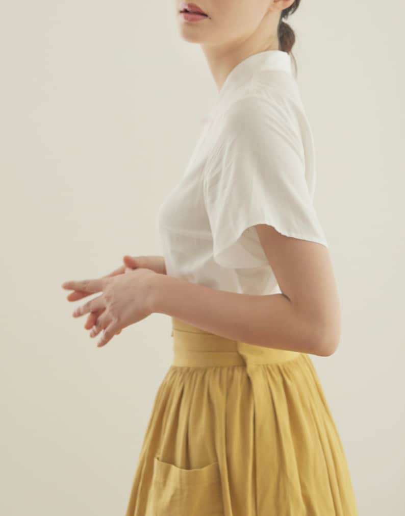 May include: A woman wearing a white short-sleeved blouse and a yellow midi skirt with a pocket. The skirt has a wide waistband.