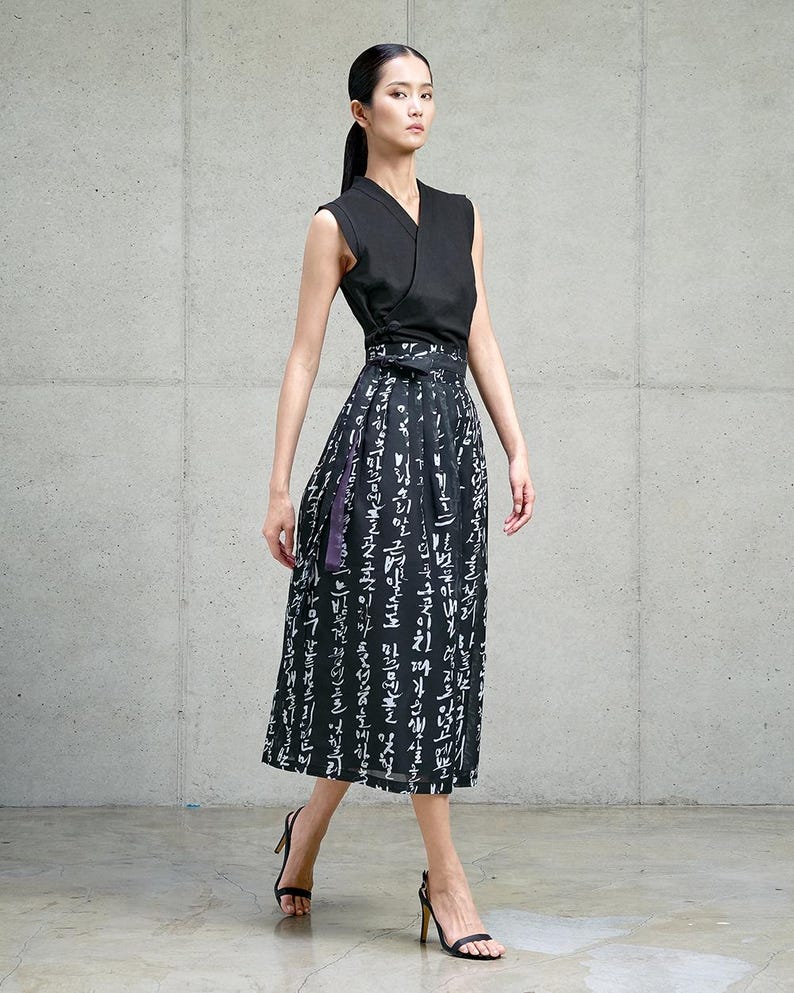 May include: A sleeveless black wrap top with a V-neckline paired with a high-waisted midi skirt. The skirt is black with a white printed pattern of Korean script characters and features a matching fabric tie belt at the waist.