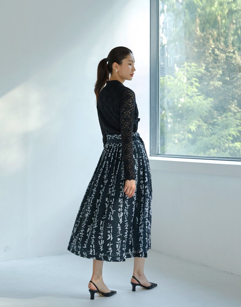 May include: A black long-sleeved lace top paired with a black midi skirt featuring a white calligraphic print. The skirt has a high waist with a tie belt and a pleated design. The ensemble is completed with black slingback heels with a low heel.