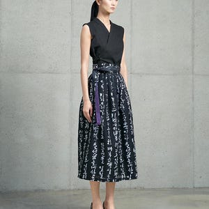 May include: A sleeveless black wrap top paired with a high-waisted midi skirt featuring a black and white calligraphic print. The skirt includes a wide waistband with a tie closure and a distinct purple fabric accent hanging from the side.